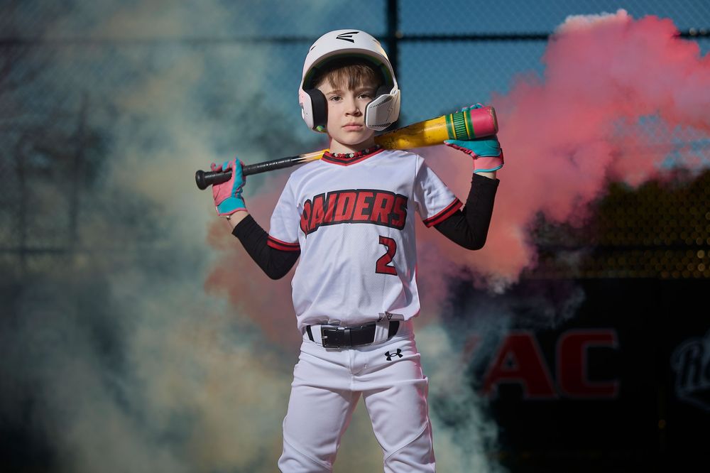 Young baseball player in Raiders uniform holding colorful bat over shoulders with pink smoke in background.