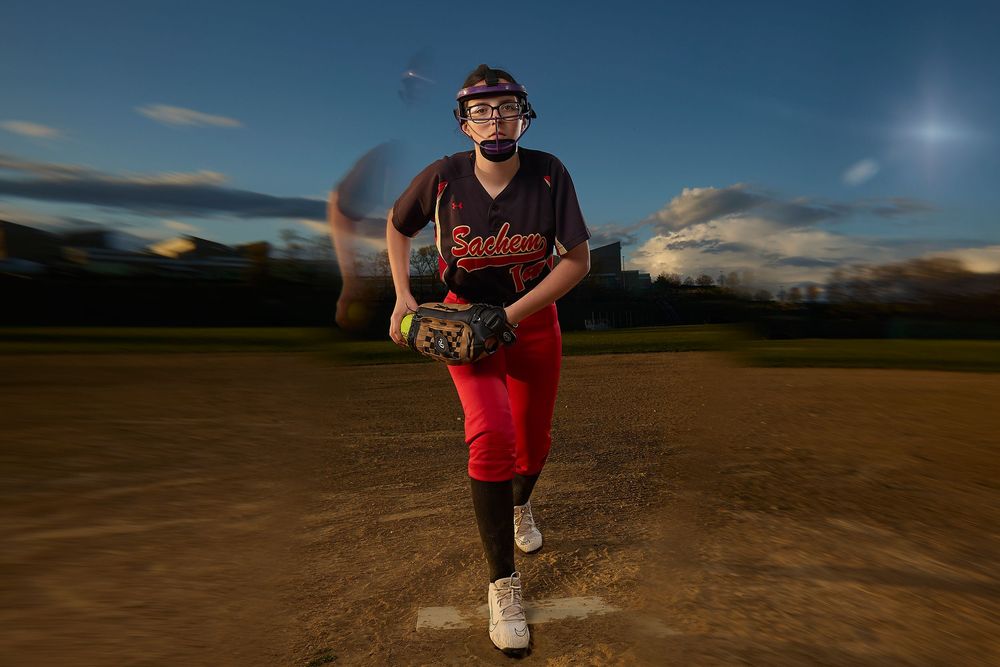 Softball pitcher in red uniform winds up on the mound at dusk under dramatic night sky lighting.