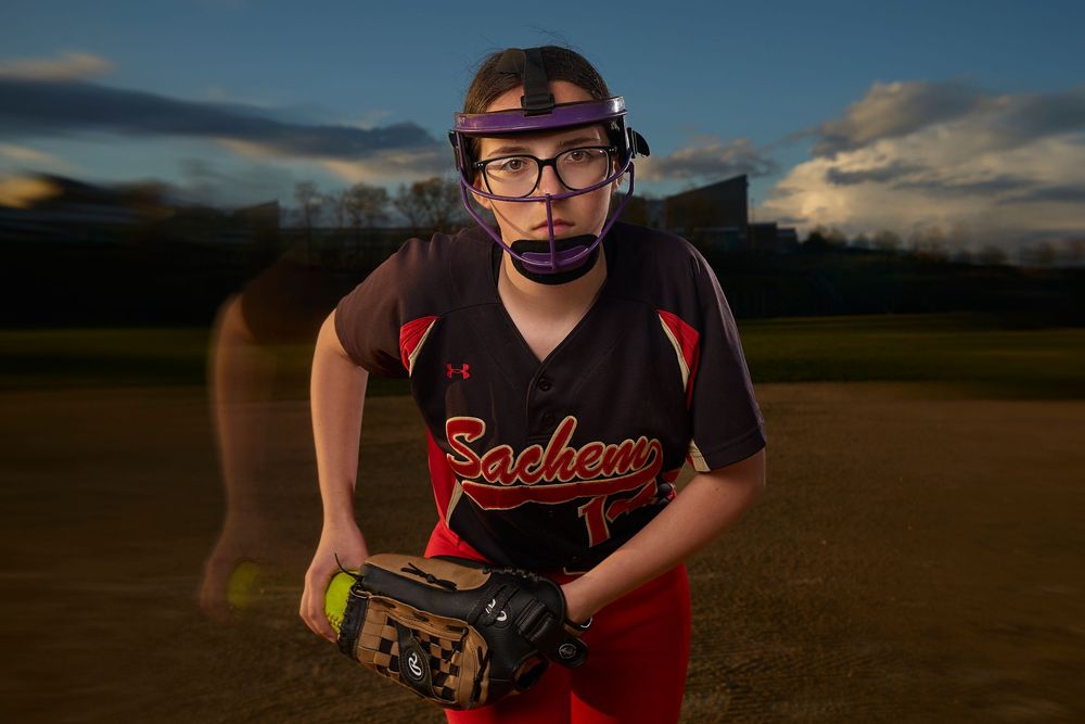 Young softball player in Sachem uniform with face guard and glove poses on field at dusk.
