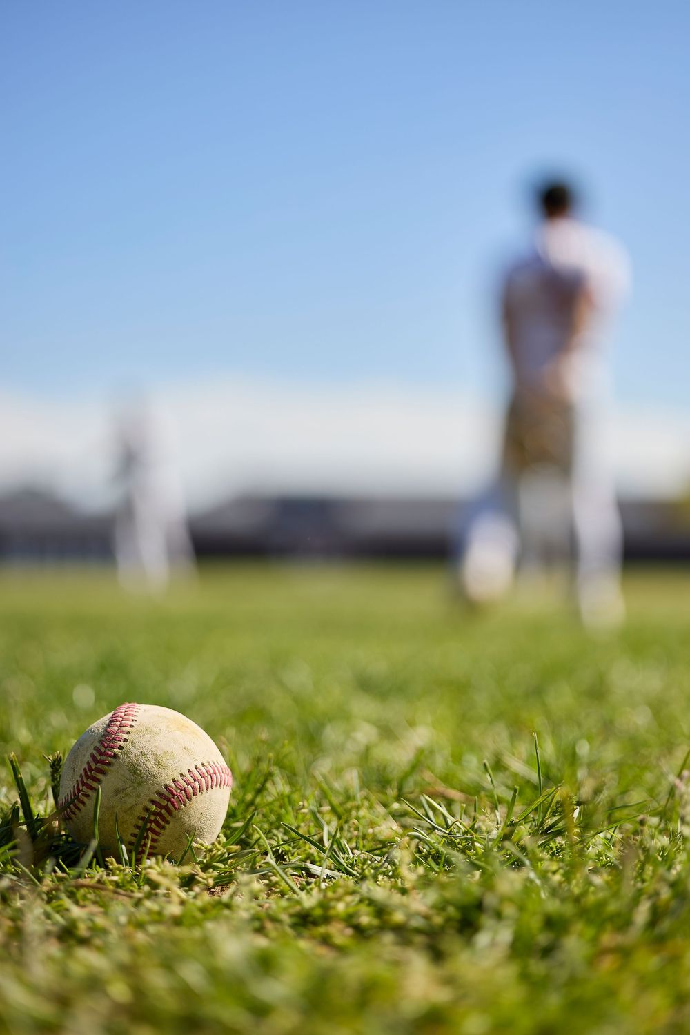 A worn baseball rests on green grass in the foreground as players practice on a sunny baseball field.