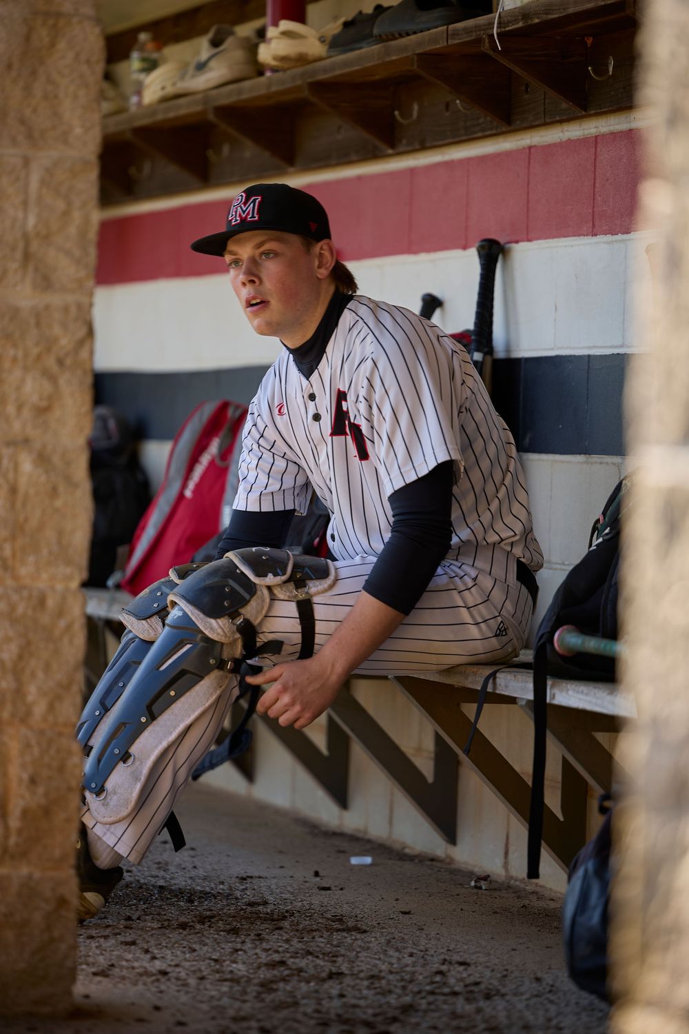 Baseball player in pinstripe uniform sitting in dugout holding glove, wearing dark cap and undershirt.