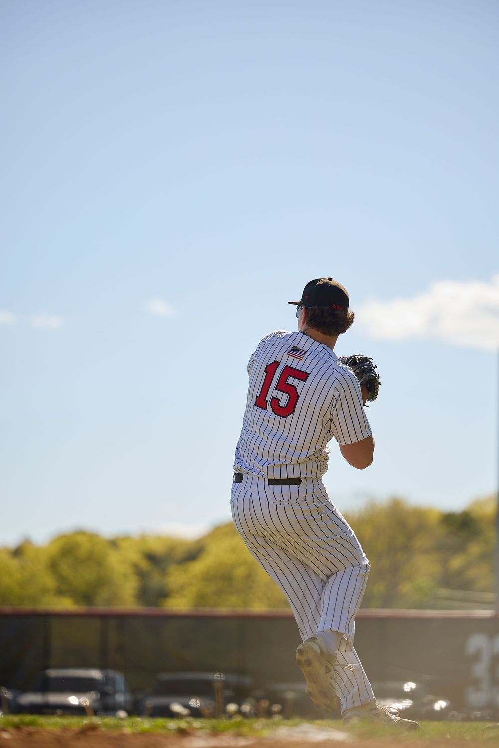 Baseball pitcher #15 in white pinstripe uniform winds up to throw during an outdoor game on a sunny day.