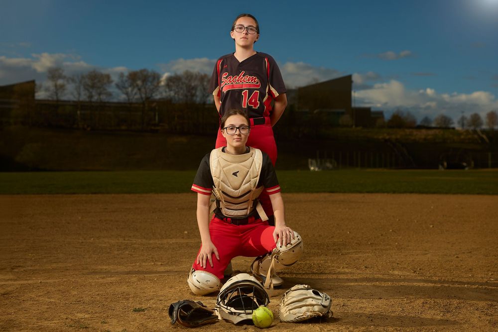 Two female softball players pose on field, catcher kneeling and pitcher standing behind.