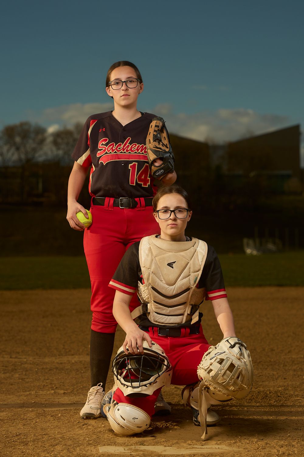 Two Sachem softball players pose on dirt field, pitcher holding ball and catcher holding helmet.