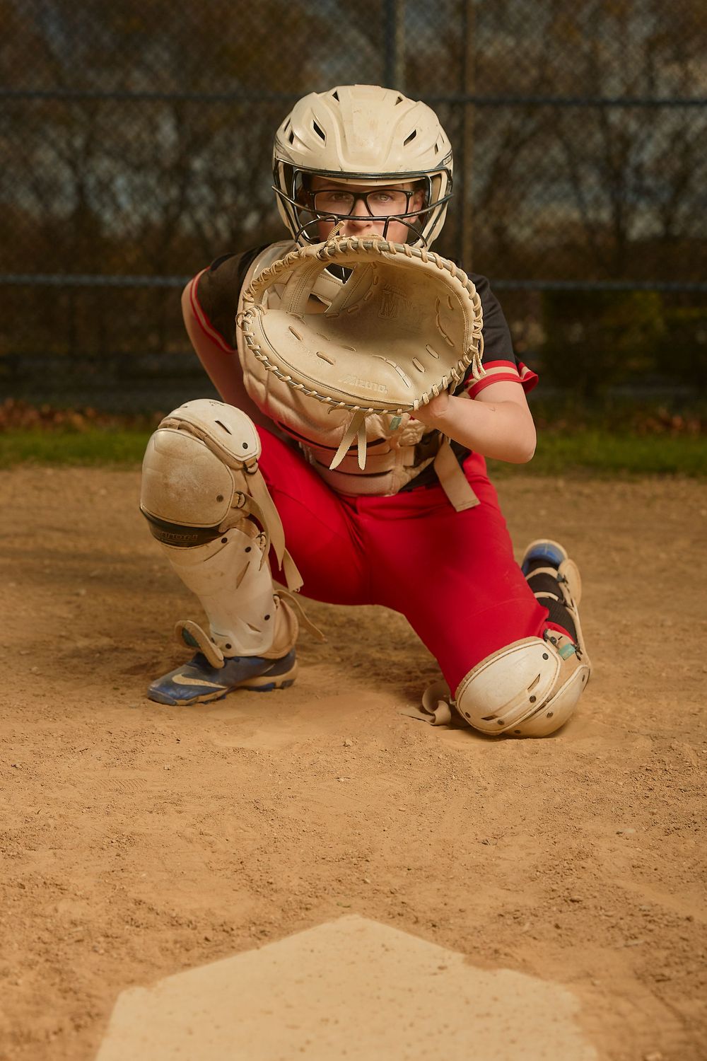 Female softball catcher in red uniform crouching at home plate with mitt raised, wearing helmet and protective gear.