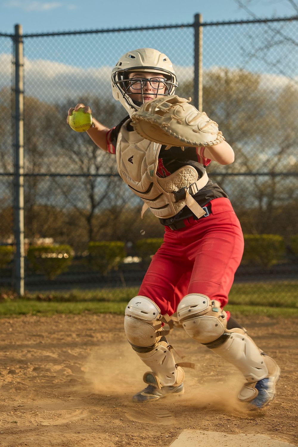Female softball catcher in red pants and full gear catching a yellow ball in a cloud of dust.