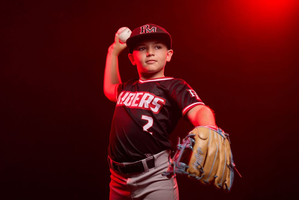 Young baseball player in Raiders uniform poses mid-throw against dramatic red backdrop.