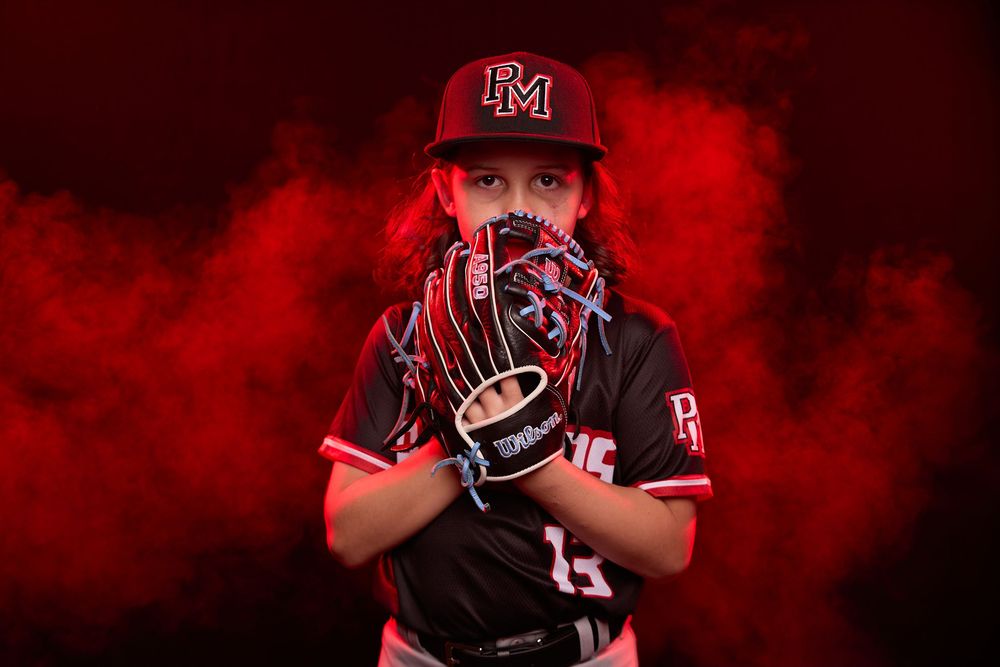 Young baseball player in PM uniform holds Wilson glove up to face against dramatic red smoke background.