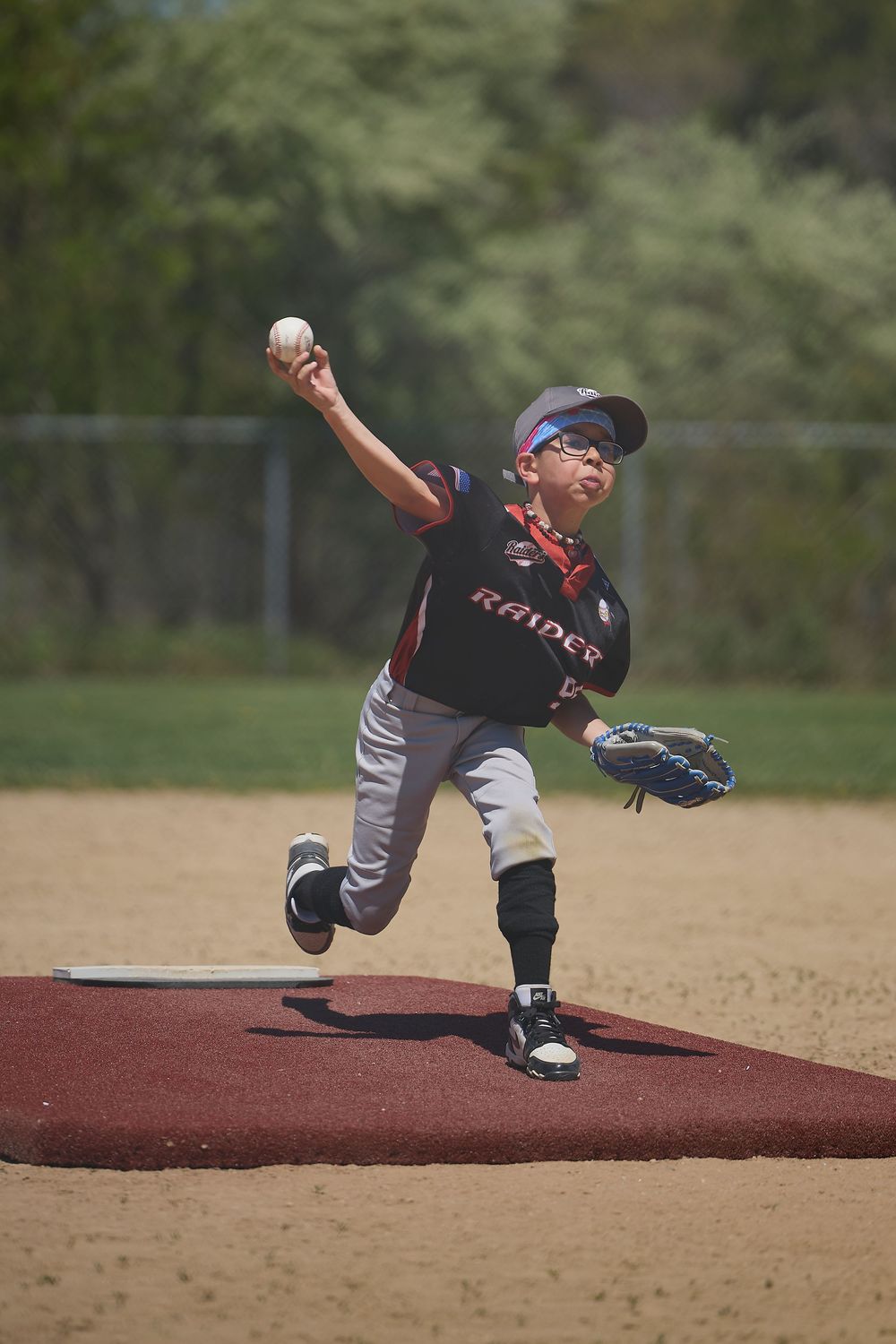 Young baseball pitcher in Raiders uniform mid-throw on pitching mound during youth league game.