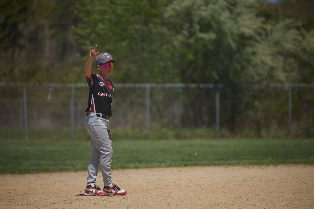 Baseball player in black uniform standing on field with one arm raised, surrounded by trees and chain-link fence.