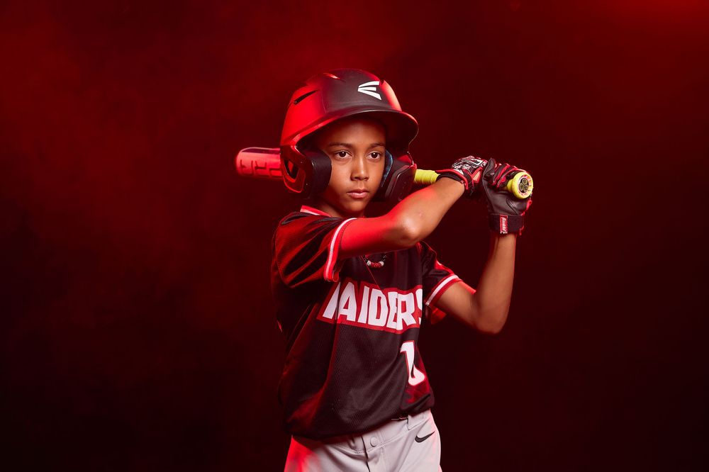Young baseball player in Raiders uniform poses with bat on shoulder under dramatic red lighting.