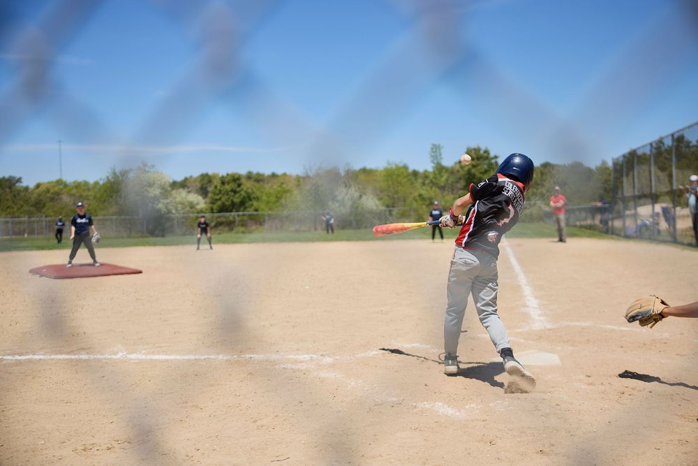 Young baseball player swings bat during a sunny little league game on a dirt field, viewed through a chain-link fence.