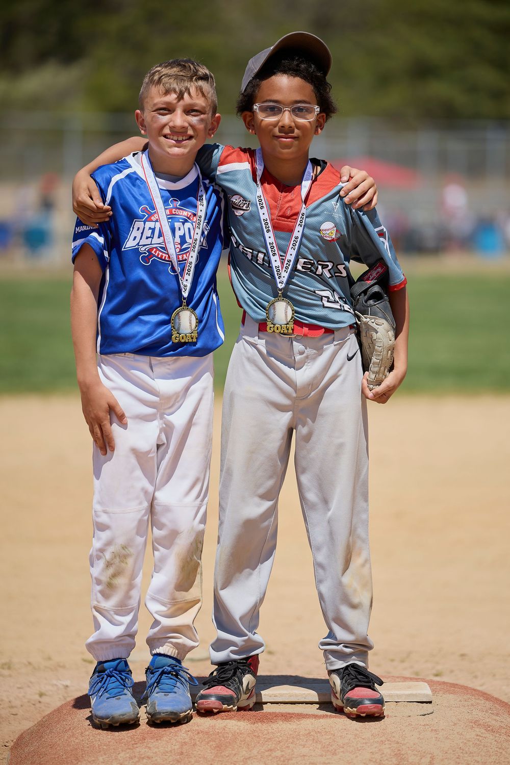 Two youth baseball players wearing medals pose together on a pitching mound after a tournament game.
