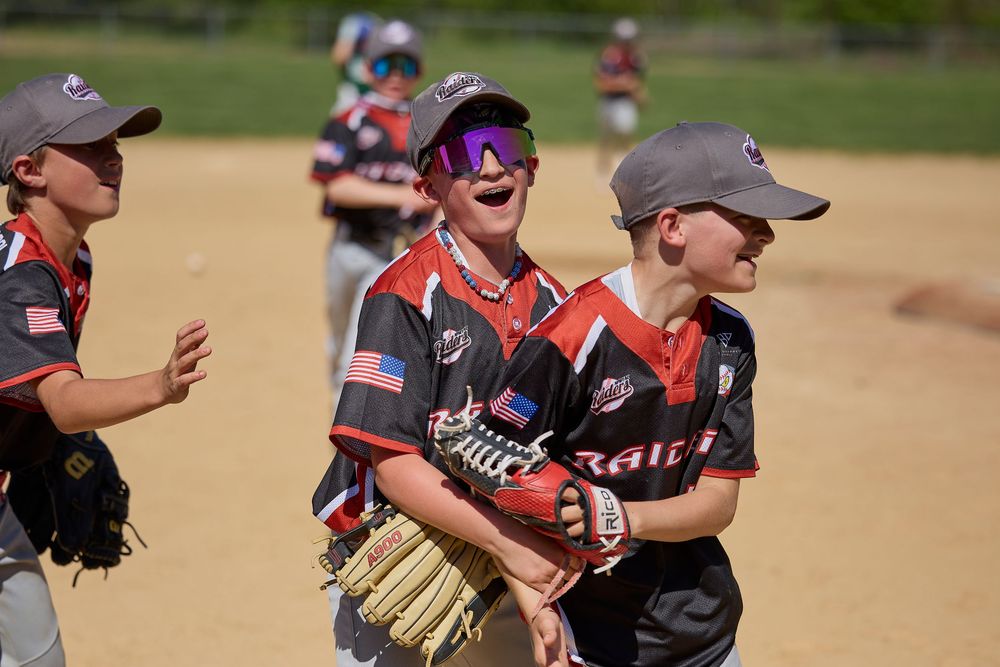 Youth baseball players in black uniforms laughing and celebrating together on a sunny baseball field.