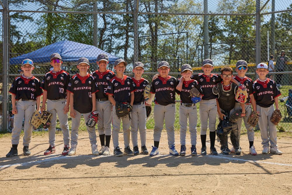 Youth baseball team posing together on a dirt field in matching dark uniforms on a sunny day.