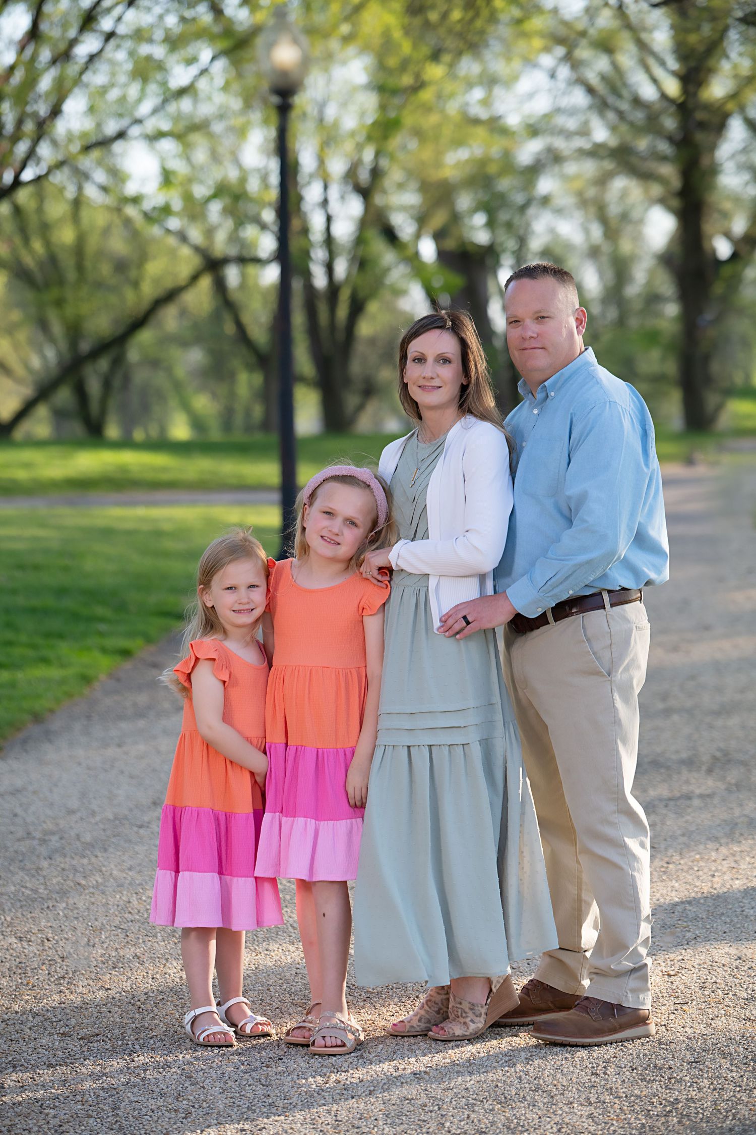 Family wearing coordinated outfits poses together on a tree-lined path in a park setting.