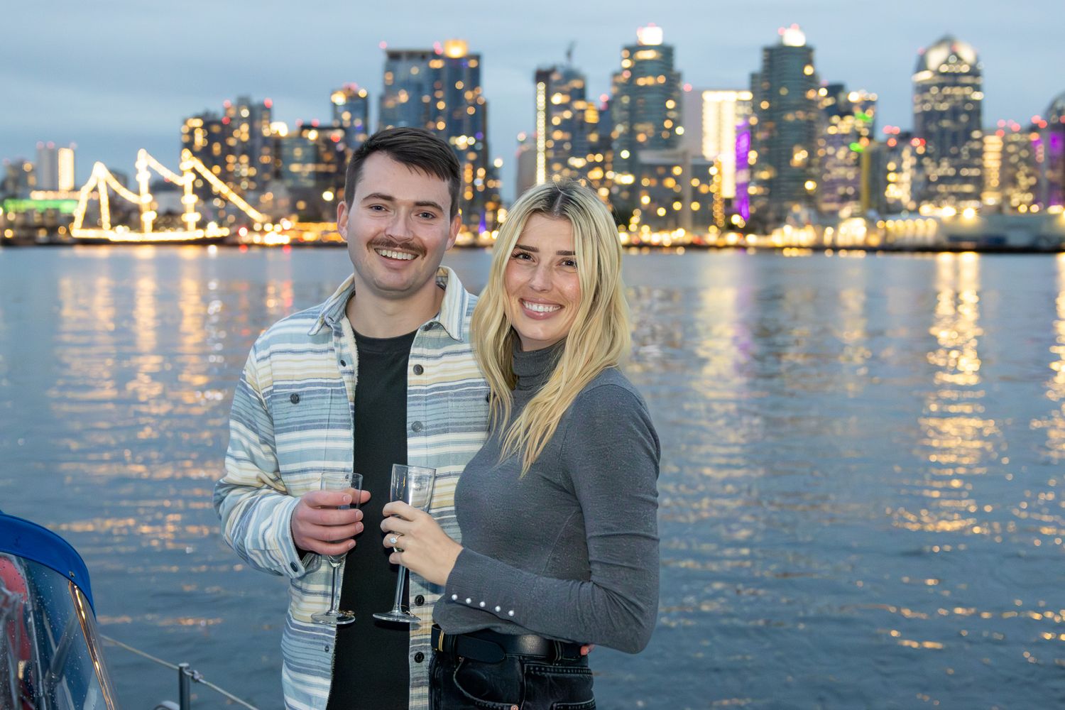 A newly engaged couple celebrating on a sailboat proposal cruise with a view of the downtown San Diego city skyline at night.