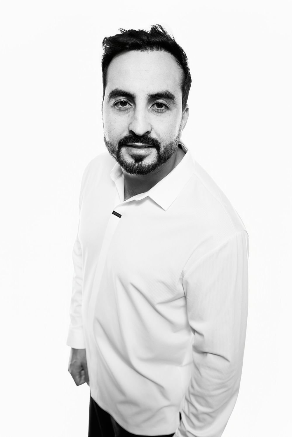 Black and white portrait of a bearded man in a white dress shirt smiling against a white background.