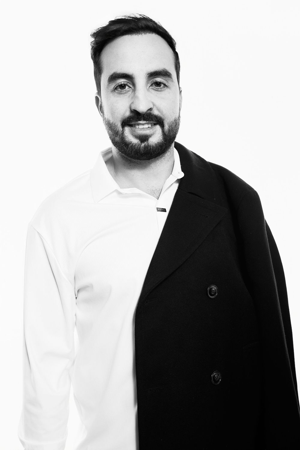 Black and white portrait of a smiling bearded man wearing a white shirt and black coat against a light background.