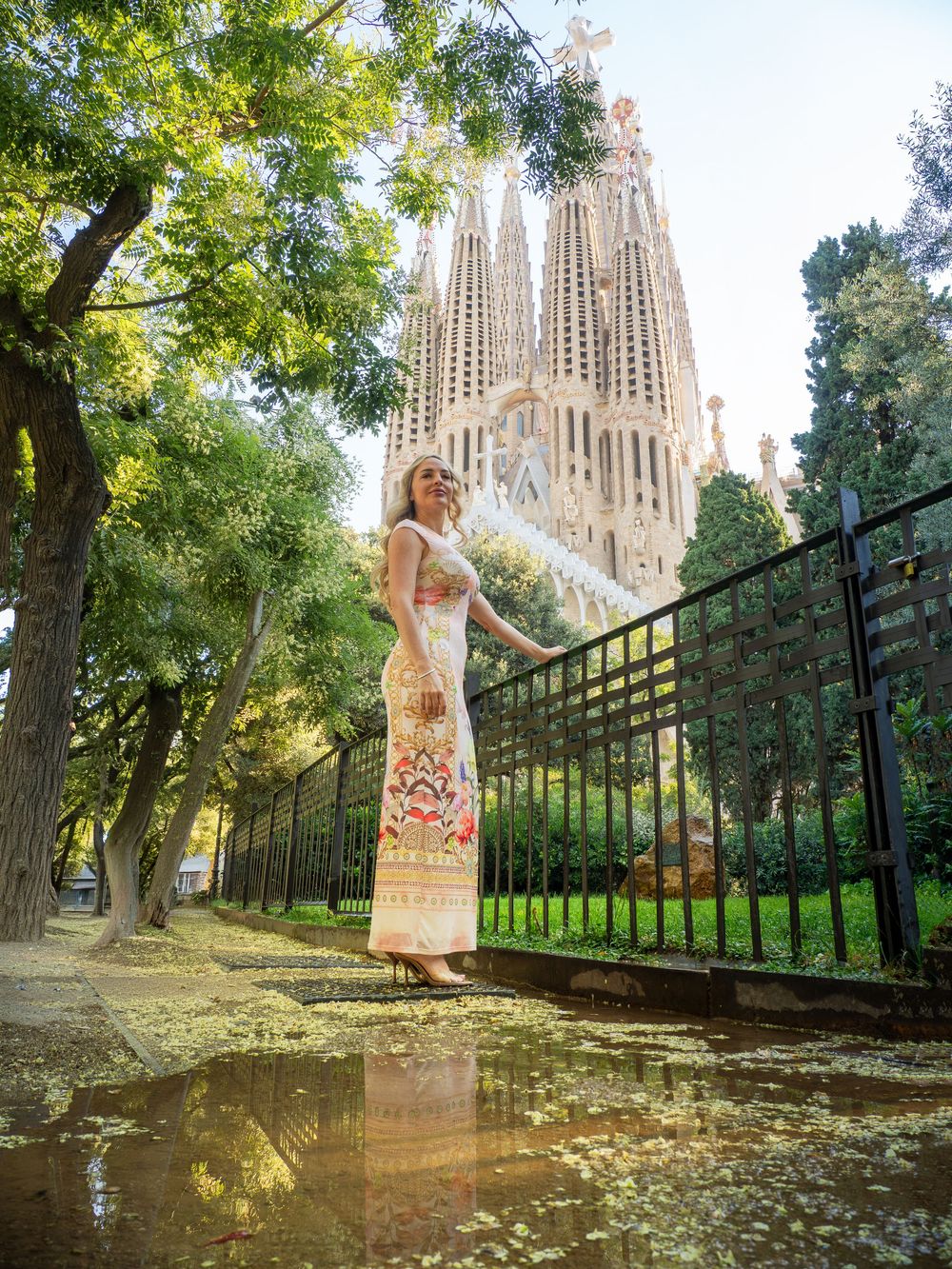 Woman in floral maxi dress posing near Sagrada Familia basilica in Barcelona, Spain, surrounded by lush trees.
