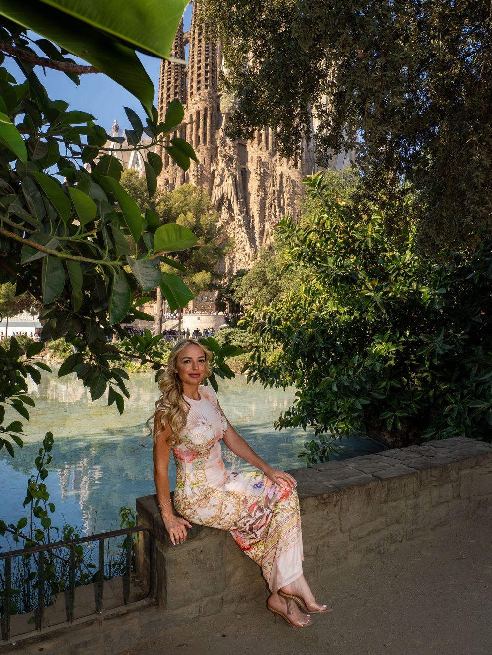 Woman in floral dress sitting by a pond with Sagrada Familia basilica and lush greenery in Barcelona, Spain.