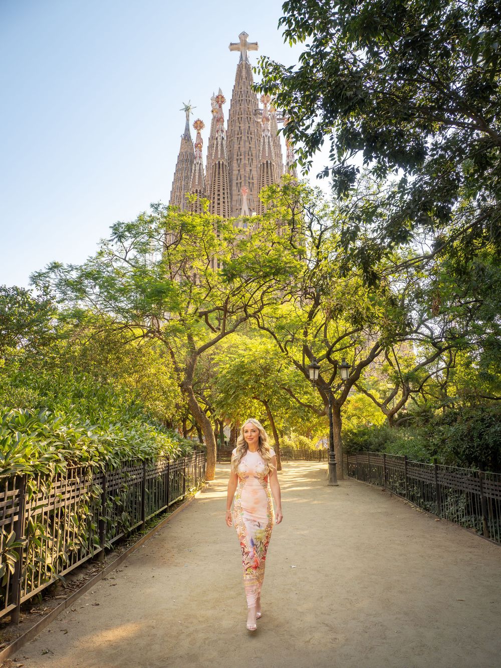 Woman in floral dress walking path before Sagrada Familia basilica, Barcelona, surrounded by lush green trees.