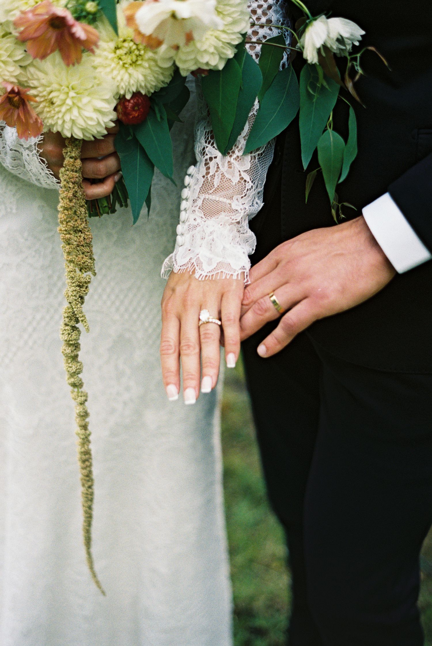 Bride in lace gown holding hands with groom in black tuxedo walking on green grass at outdoor wedding ceremony.