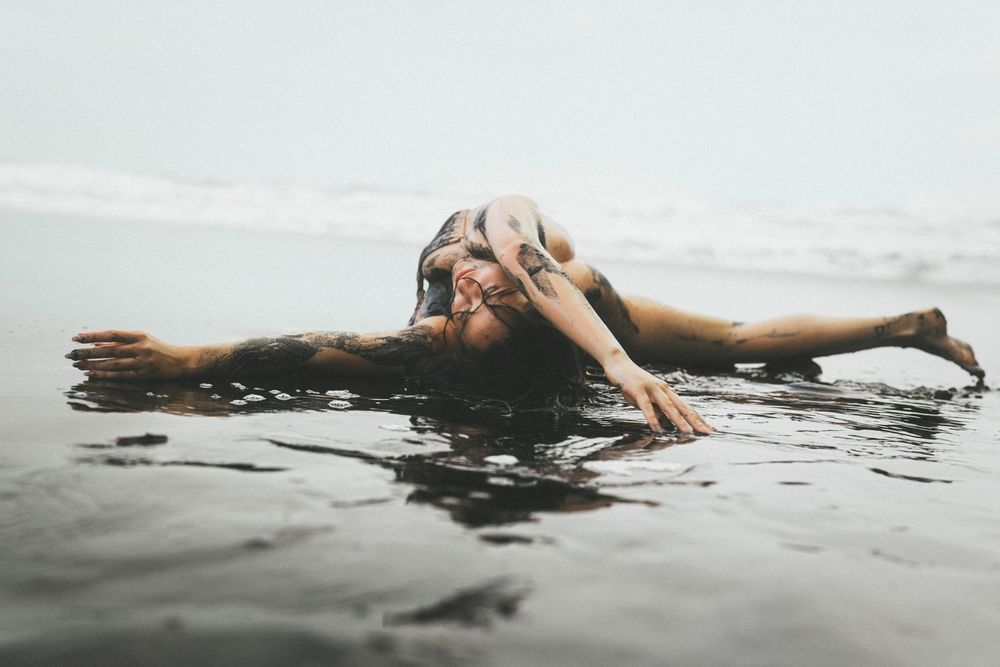 A tattooed woman lies gracefully in shallow ocean water, arms outstretched, with waves gently surrounding her.