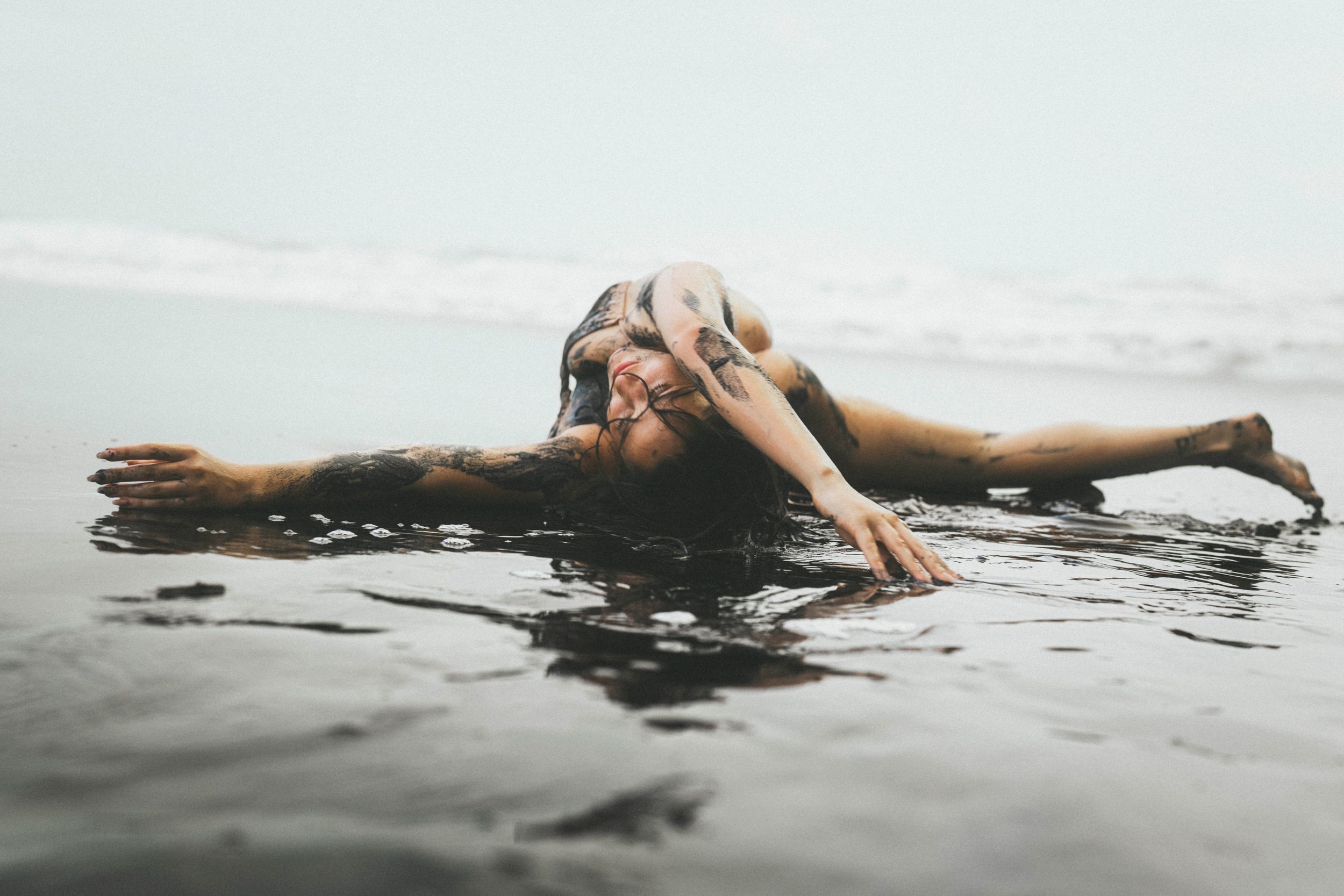 A tattooed woman lies gracefully in shallow ocean water, arms outstretched, with waves gently surrounding her.