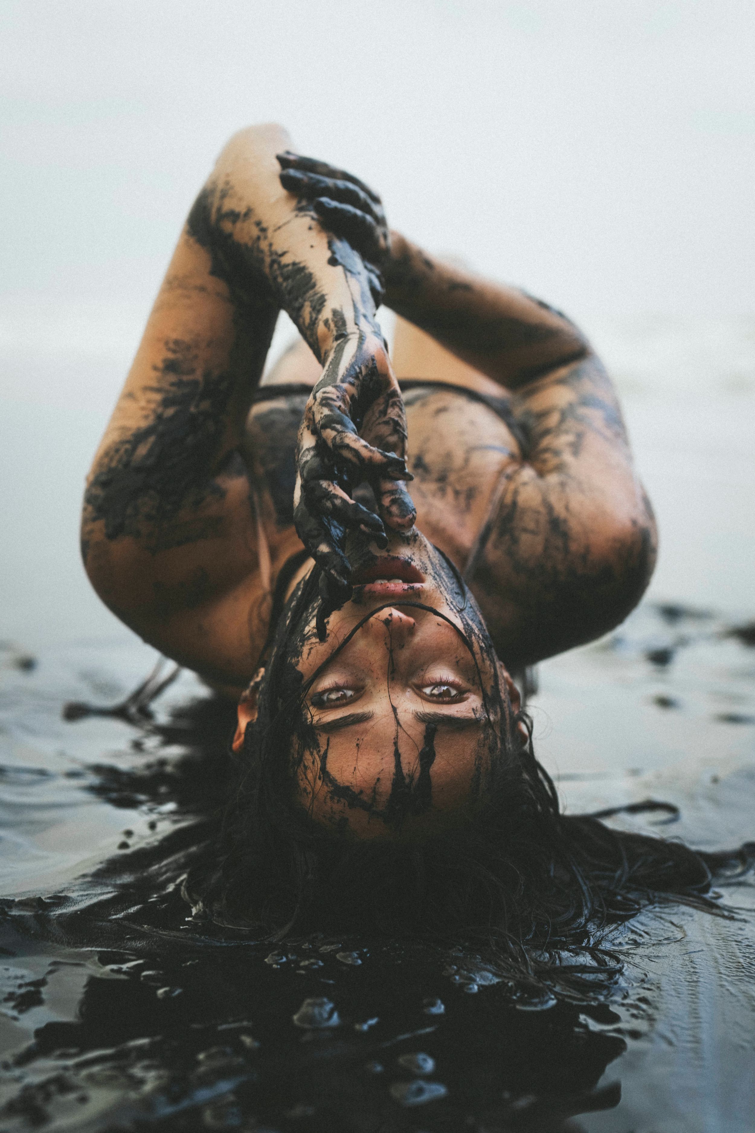Person covered in dark mud lying in water with hair raised, artistic mud bath photography.