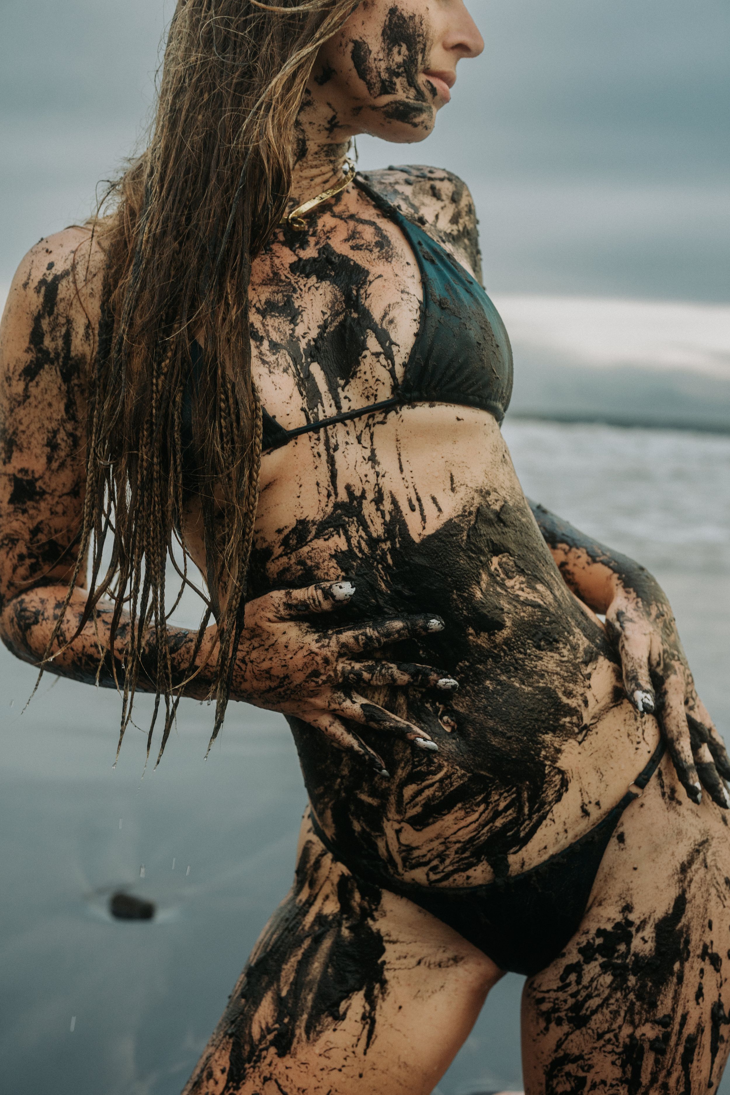 Woman in bikini covered in dark mud posing on a beach with overcast skies and ocean waves behind her.