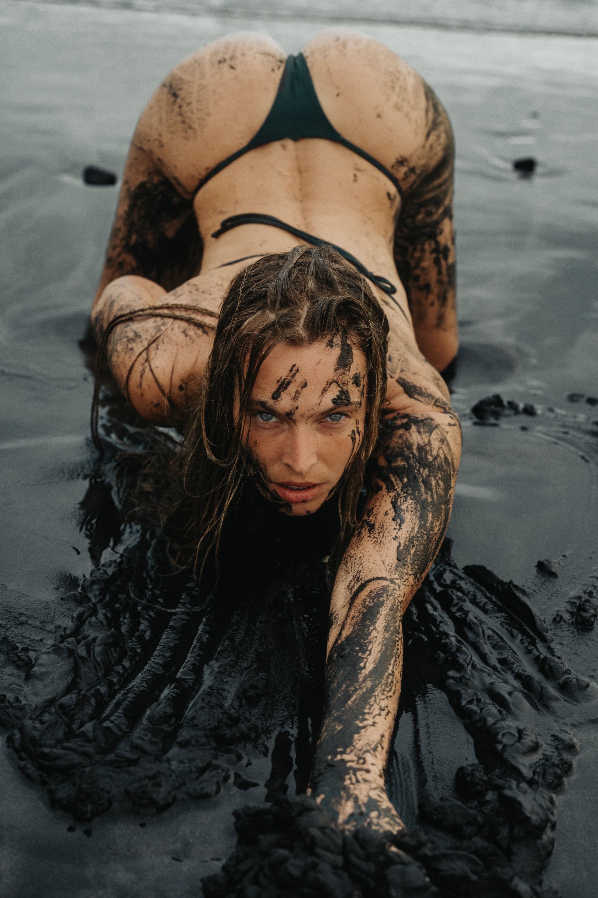 Woman covered in dark mud posing on a black sand beach wearing a green bikini.