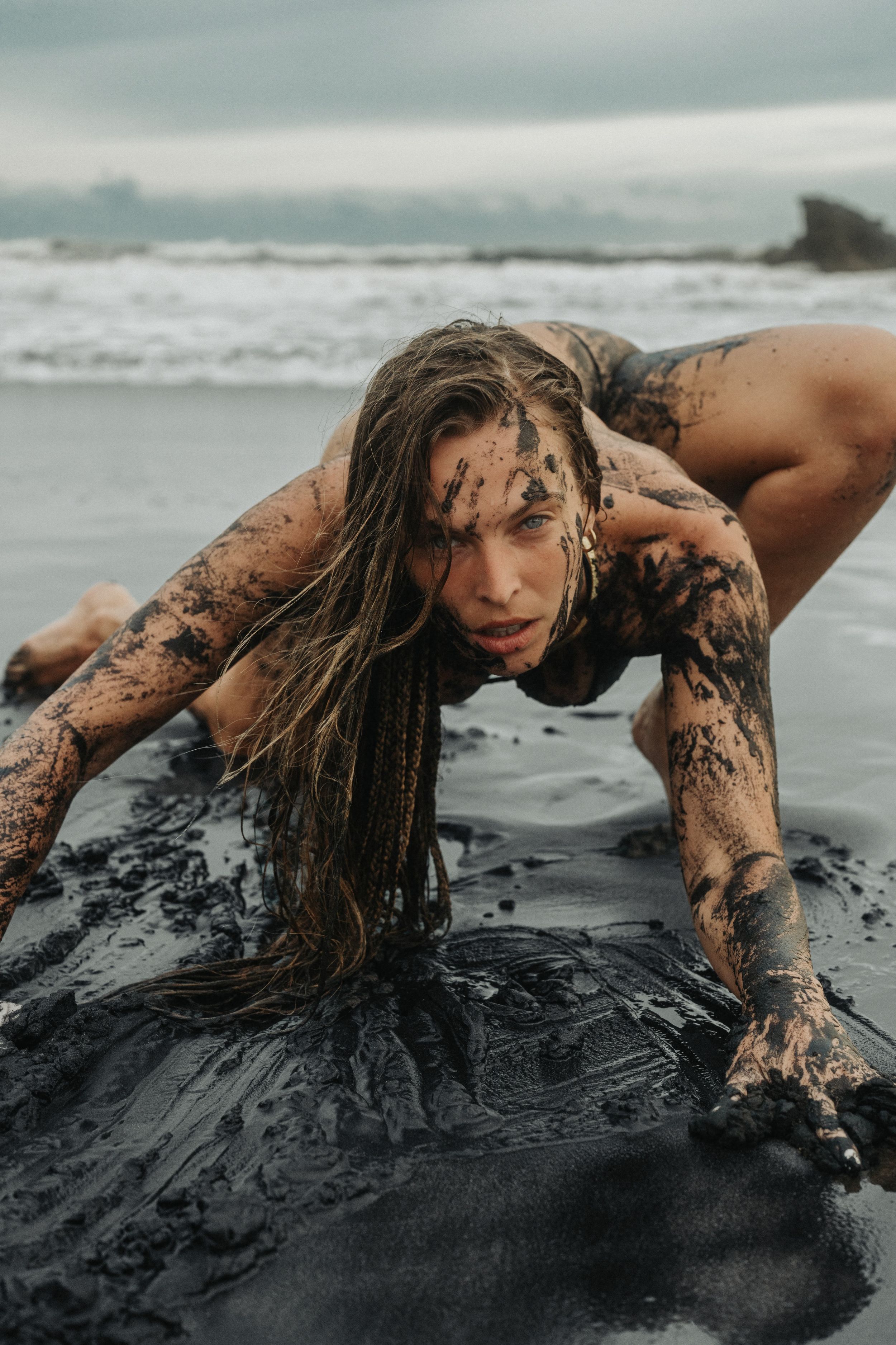 Woman covered in black sand and mud posing dramatically on a dark beach with ocean waves in the background.