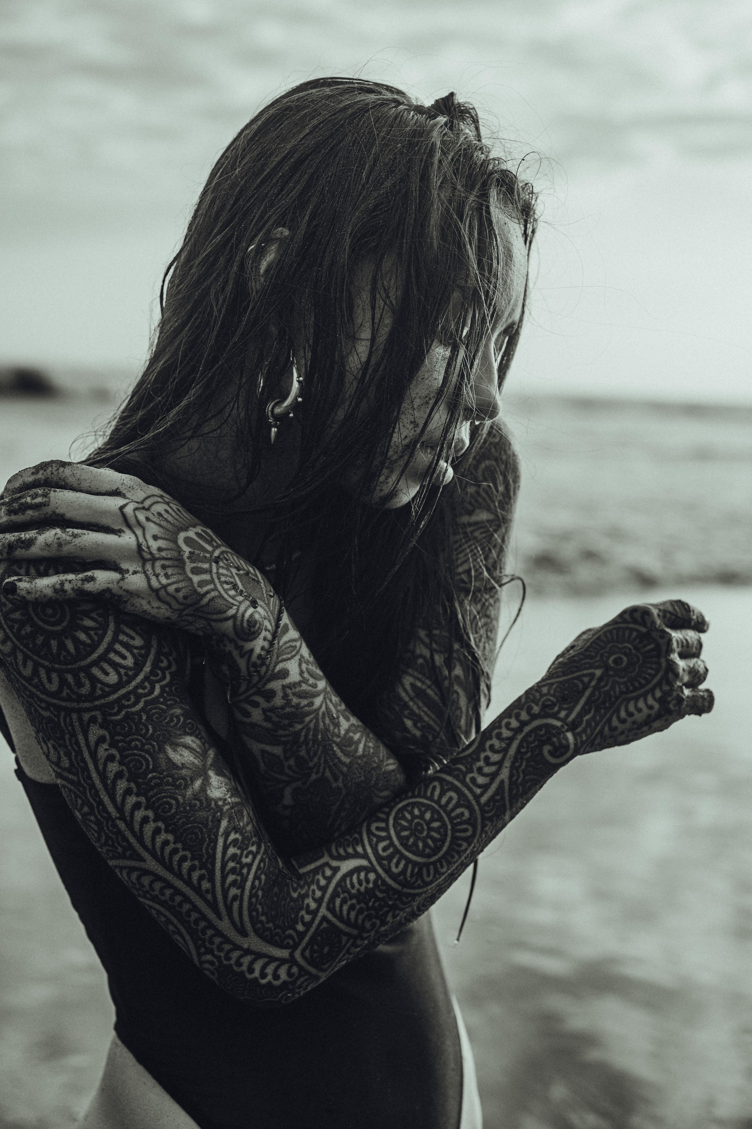 Black and white photo of a tattooed woman with long hair at the beach, arms crossed over her face.