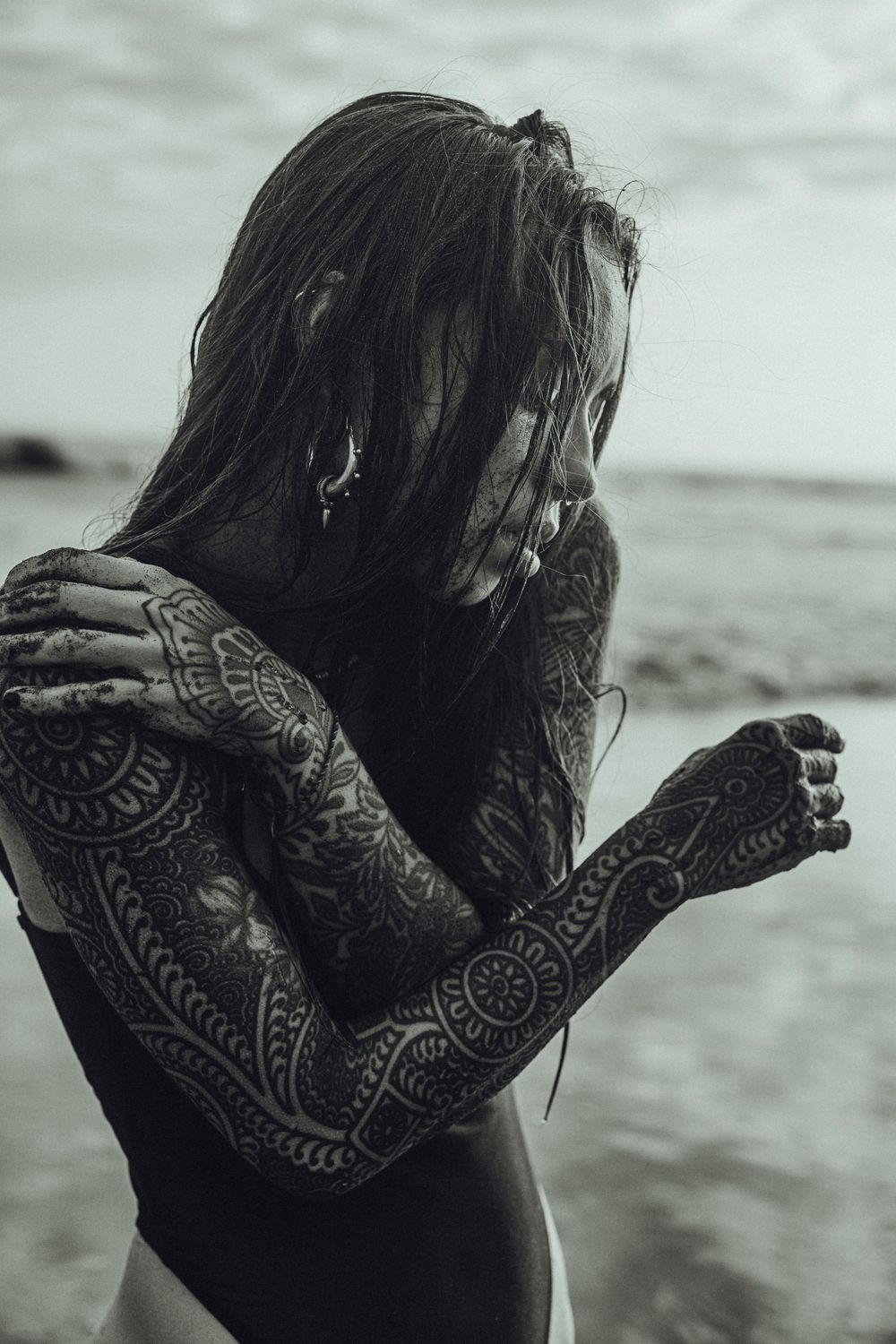 Black and white photo of a tattooed woman with long hair at the beach, arms crossed over her face.