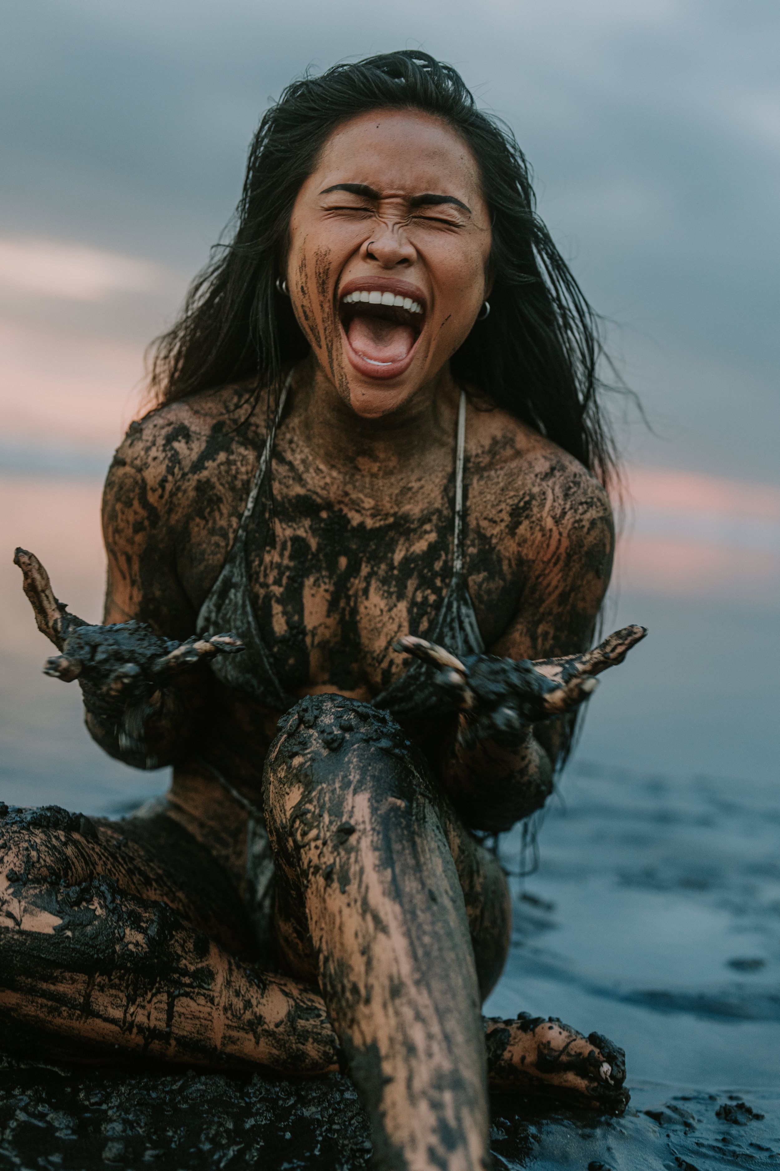 Woman covered in dark mud sitting by the ocean, laughing with mouth wide open in a joyful expression.