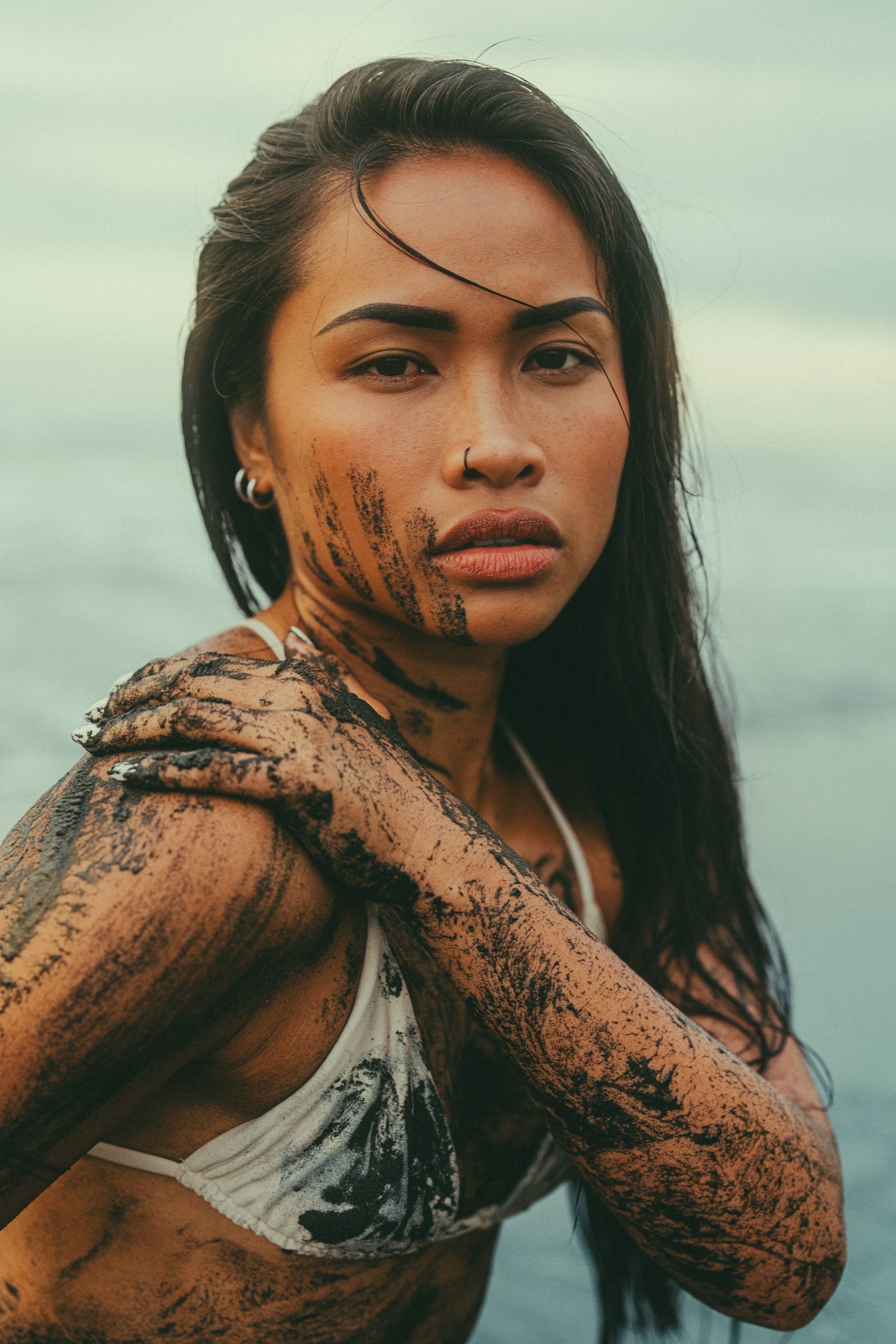 Young woman with mud on her face and arms posing outdoors near water in a white bikini top.