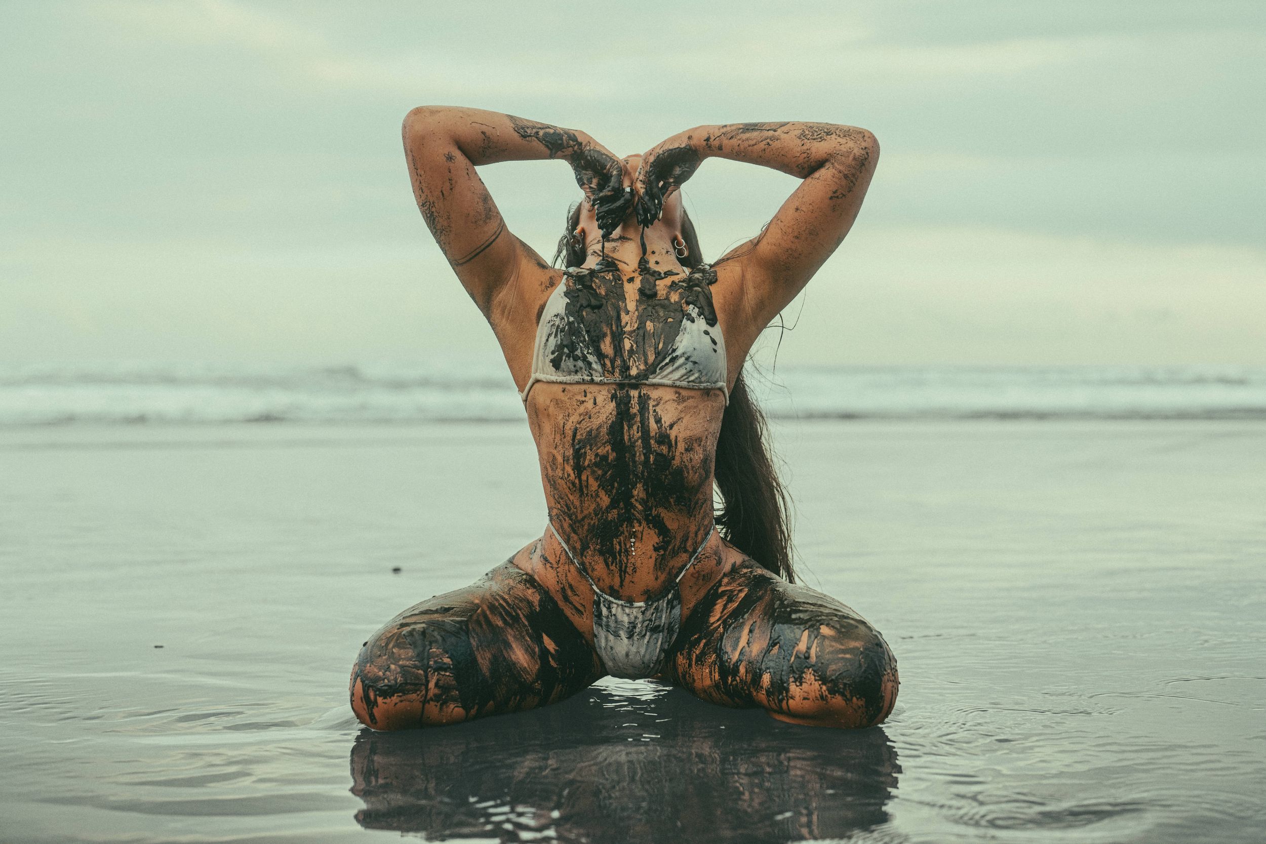 Woman covered in dark mud sits on wet beach shoreline in yoga pose with hands behind head.