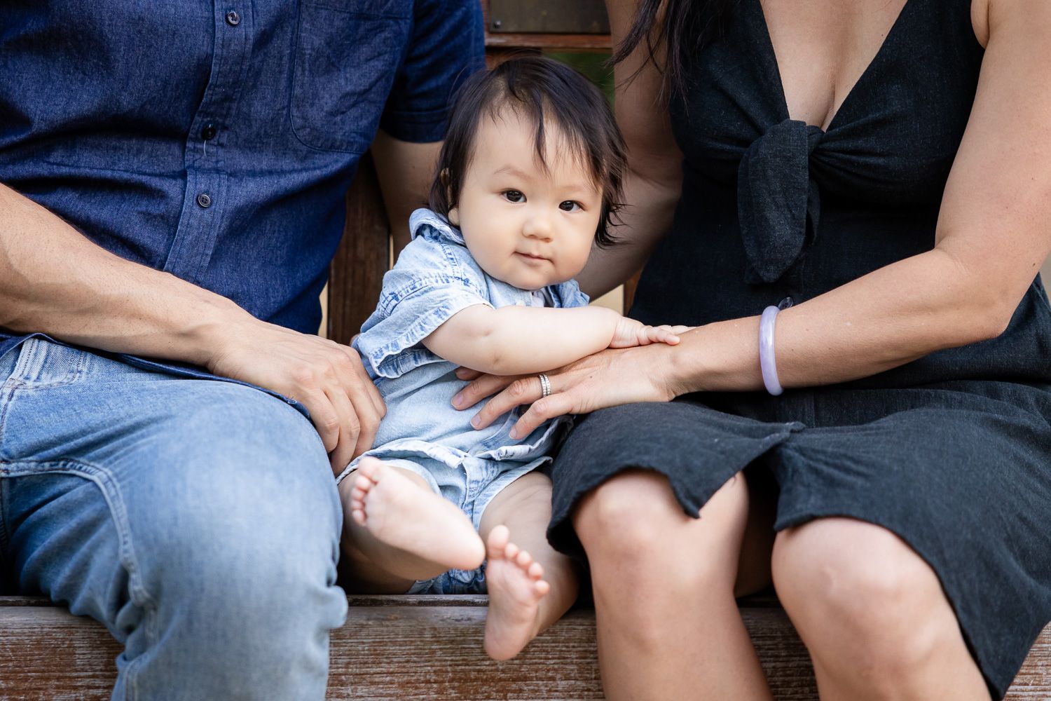 Baby sitting between parents on a bench, wearing a blue romper with bare feet in a family portrait.