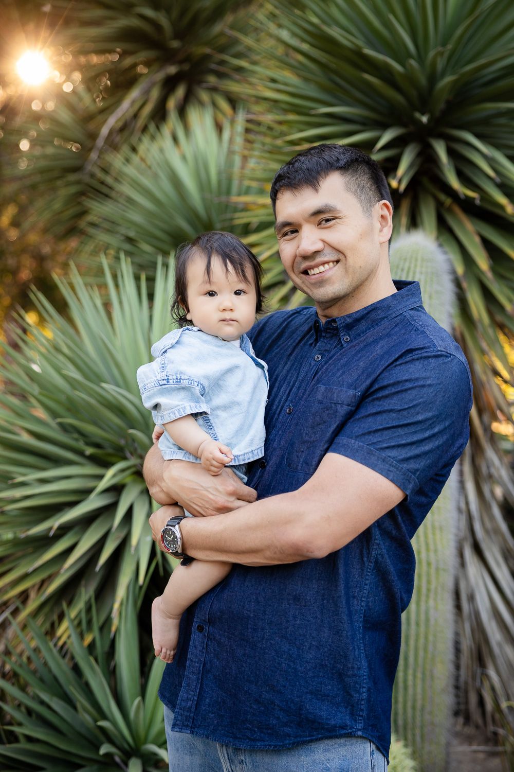 Smiling dad holding a baby, posing outdoors among green cacti for their family photography session in Palo Alto