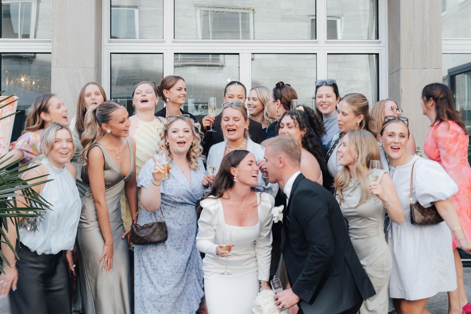 Bride and groom kissing surrounded by laughing wedding guests in elegant attire outside a modern building.