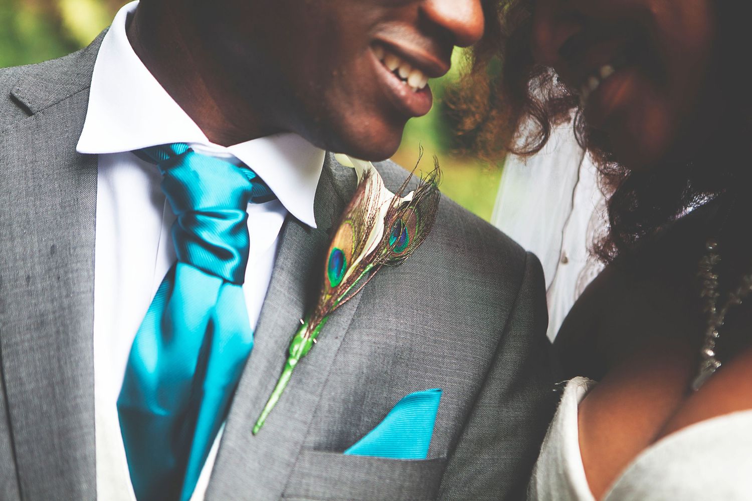 Groom in grey suit with teal tie and peacock feather boutonniere smiles with bride at wedding ceremony, at Oaks farm in Surrey