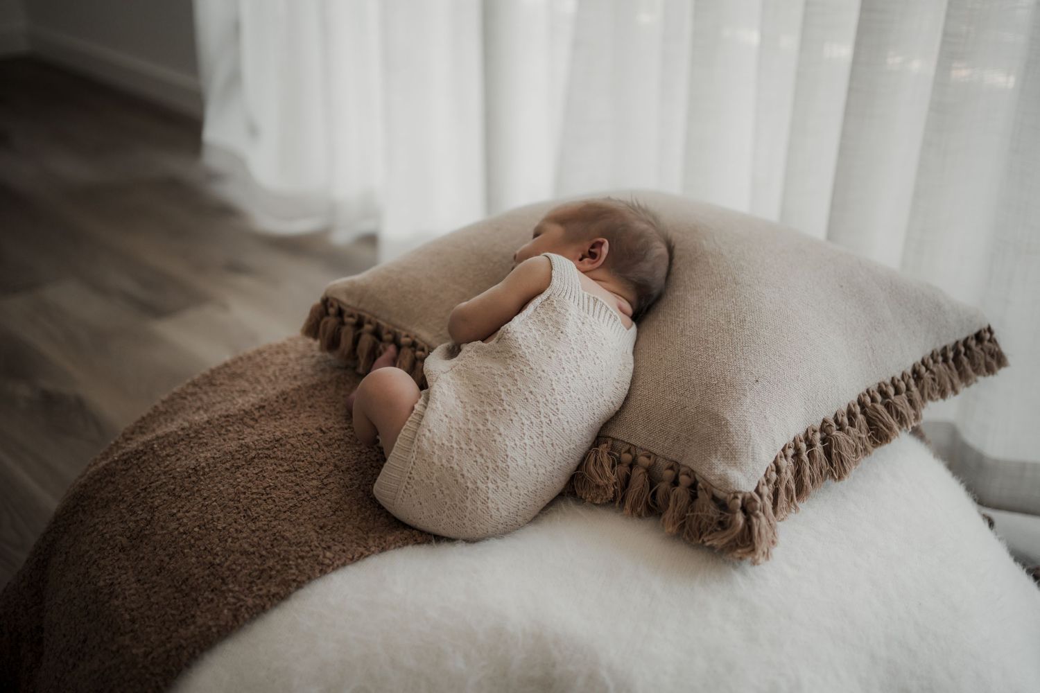 Newborn baby sleeping peacefully on a fluffy white cushion with a beige fringe pillow and soft brown blanket in the Buxton Studio