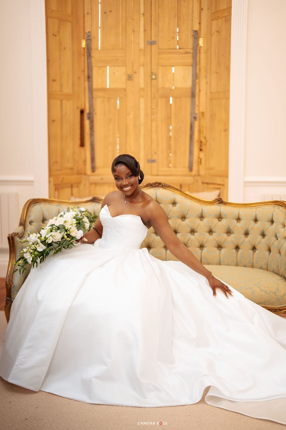 Smiling bride in a strapless white ball gown holding a white bouquet, seated on a tufted vintage sofa.
