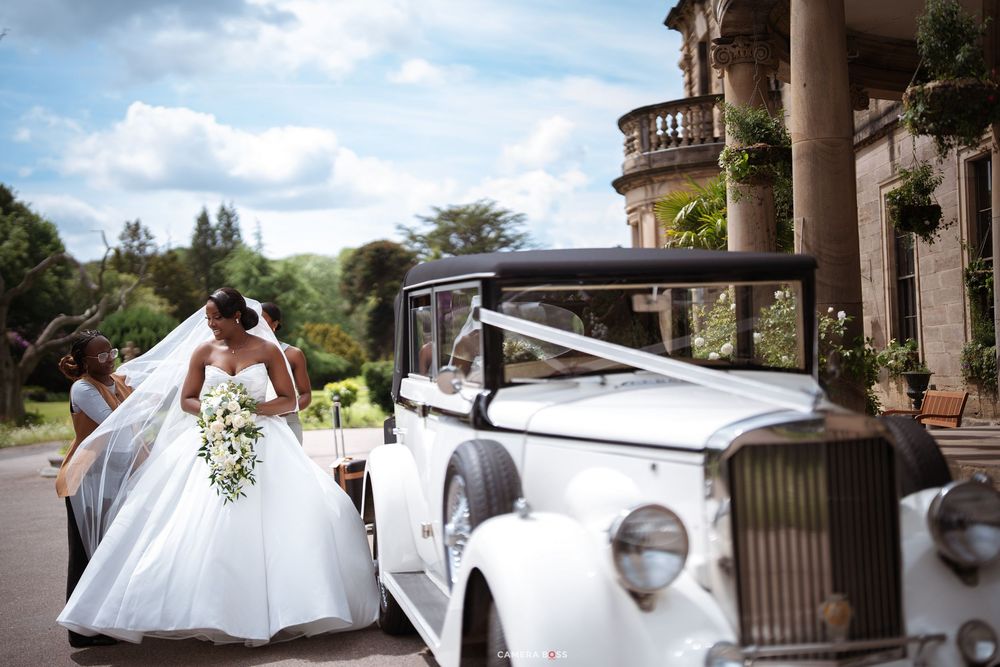 Bride in white gown with veil stands beside vintage white wedding car outside elegant manor venue.