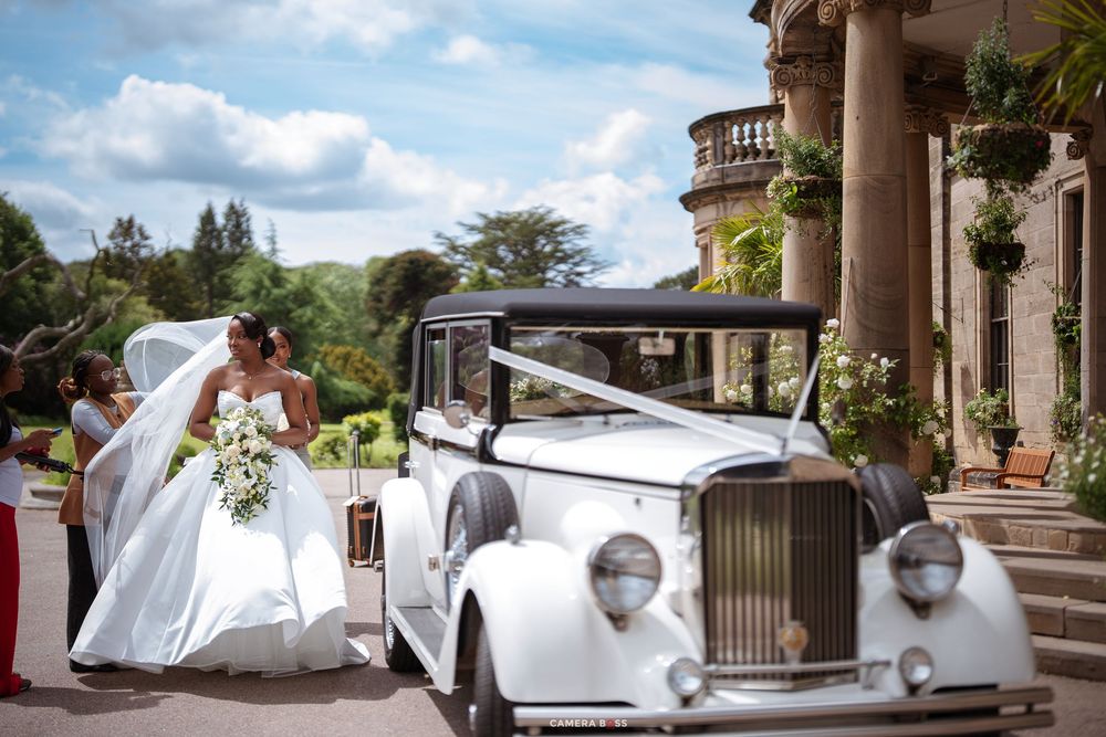 Bride in white gown beside vintage Rolls-Royce at elegant manor wedding venue with lush gardens.