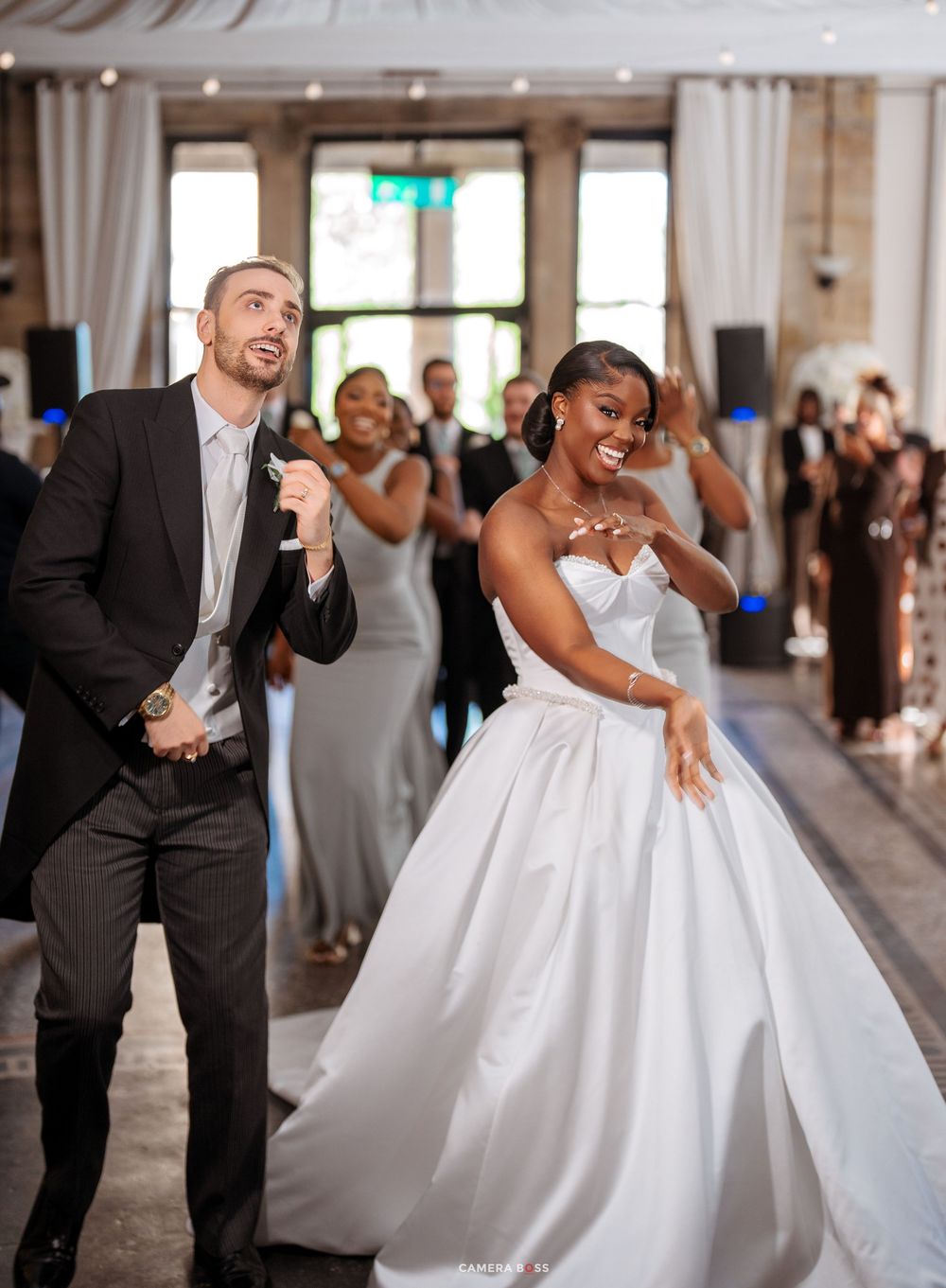 Joyful interracial newlywed couple dancing at wedding reception, bride in white ballgown, groom in dark suit.