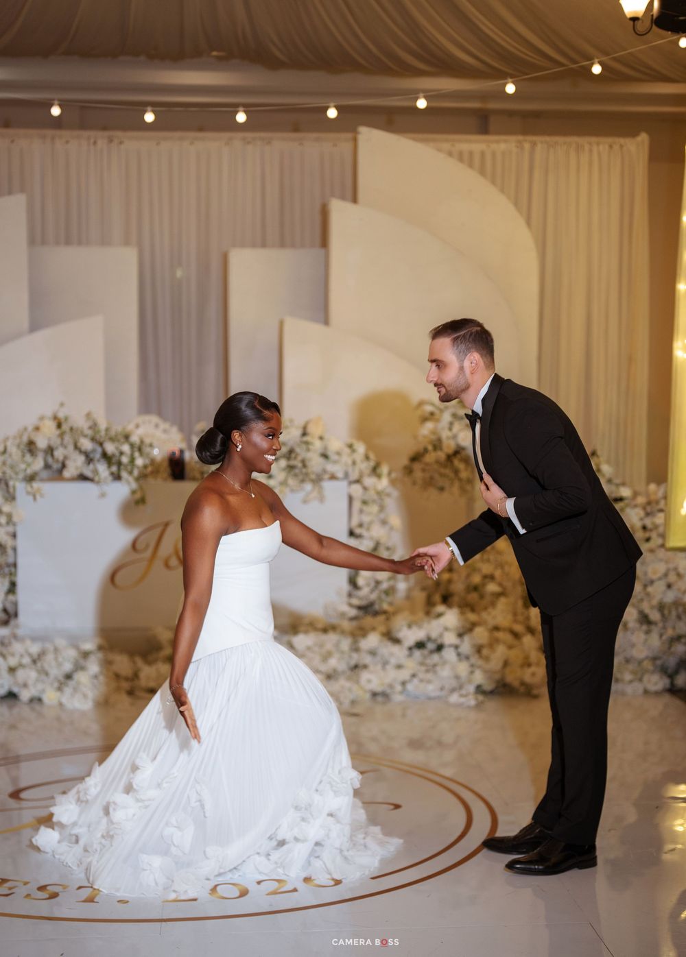 Interracial bride and groom sharing first dance, elegant white floral backdrop, bride in flowing white gown.