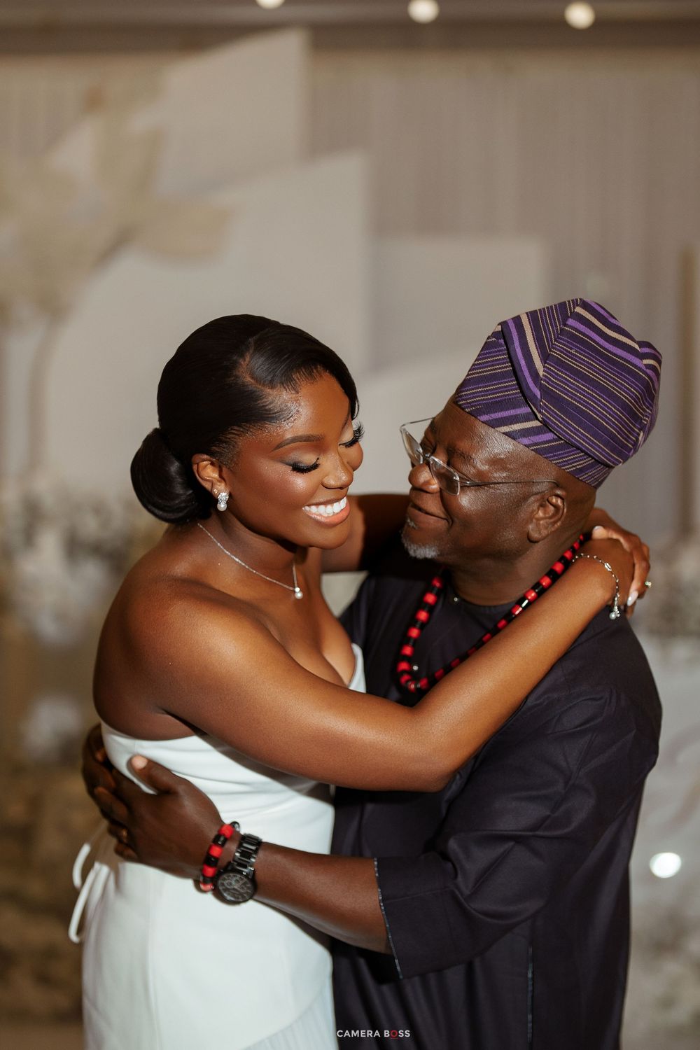 A bride in a white gown shares a joyful embrace with an elder man in traditional African attire at a wedding reception.