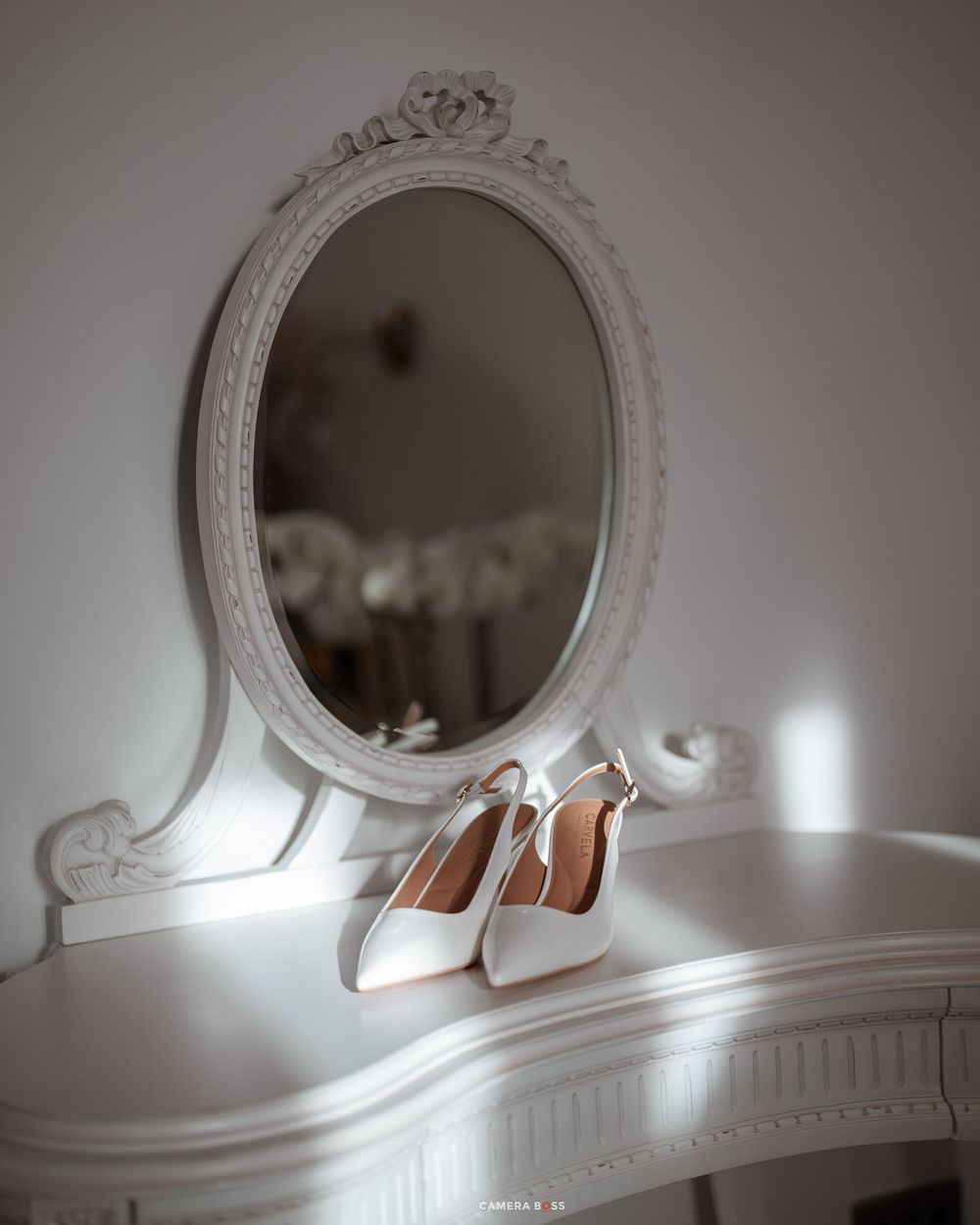 Elegant white bridal heels displayed on vintage white vanity table with ornate oval mirror.